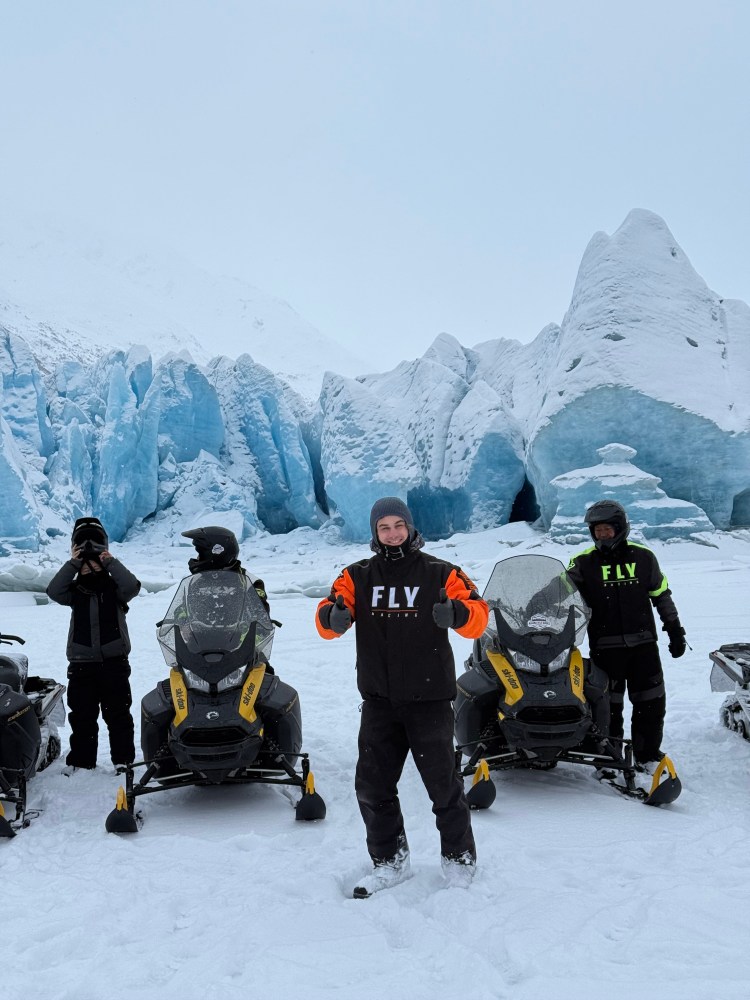 People in winter gear posing with snowmobiles in front of blue ice formations on a snowy landscape.